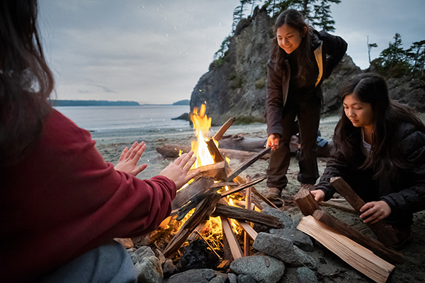Three sisters gathered around a campfire with water in the background