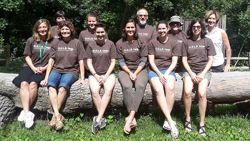 A group of eleven adults wearing FIELD Corps shirts, half sitting on a log and half standing behind them