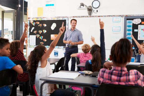 teacher standing in front of a whiteboard with a class of students raising their hands
