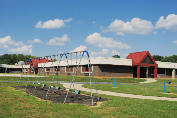 playground swings in foreground with a single-story school in the background and a blue sky with clouds